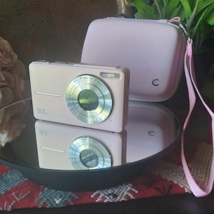 May include: A light pink digital camera with a silver lens and a matching pink protective carrying case. The camera features a wrist strap and a silver circular lens housing. The device is displayed on a reflective surface, showing its front profile.