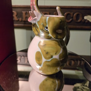 May include: A ceramic elephant creamer with a white base and olive green spots. The elephant's trunk is raised, revealing a pink interior. The creamer is reflected in a round mirror, showcasing its unique design.