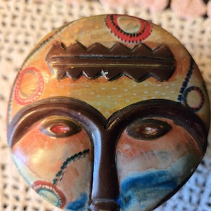 May include: A circular, painted mask with a face design. The mask features brown, orange, blue, and red colours. The eyes are red, and the mask has a brown, jagged crown-like shape at the top. The mask is likely a decorative item.