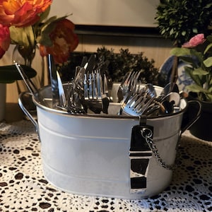 May include: A white metal utensil holder filled with silverware, including forks, knives, and spoons. The holder has two handles and a decorative tag. The holder sits on a white lace tablecloth, with flowers in the background.