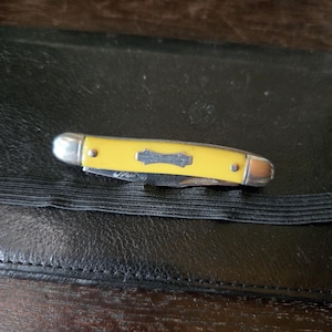 Puede incluir: Una pequeña navaja de bolsillo amarilla con extremos de metal plateado y un emblema decorativo. La navaja está cerrada y descansa sobre una superficie de cuero negro. La hoja no es visible.