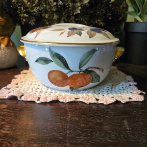 May include: A white ceramic serving bowl with a lid, featuring a gold rim and handles. The bowl is decorated with a branch of oranges and green leaves. The lid has a pattern of berries and leaves. The bowl sits on a crocheted doily.