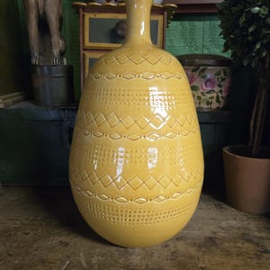 May include: A yellow ceramic vase with a textured surface and geometric patterns. The vase has a bulbous body and a slender neck. The design includes diamond and dot patterns. The vase is likely used for decorative purposes.