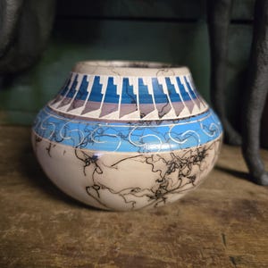 Puede incluir: Un jarrón de cerámica hecho a mano con forma redondeada. El jarrón presenta una base color crema con intrincados patrones negros en forma de relámpago. La parte superior muestra un diseño geométrico en azul, gris y blanco. Una banda azul rodea el centro.