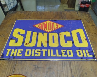 Puede incluir: Un letrero rectangular de Sunoco con fondo azul y letras amarillas. El letrero dice "SUNOCO THE DISTILLED OIL" en negrita, con una fuente de estilo vintage. El logotipo de Sunoco es un diamante amarillo con las palabras "SUN OILS" en rojo.