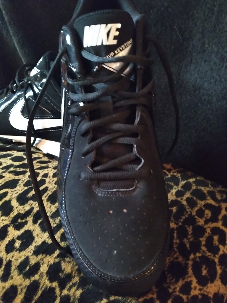 May include: Black Nike athletic shoes with white laces and a white Nike logo on the side. The shoes have a black suede toe and a black rubber sole.