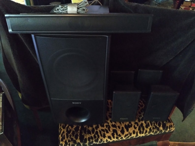 Puede incluir: Un sistema de cine en casa Sony negro con una barra de sonido, un subwoofer y cuatro altavoces sat&eacute;lite. El subwoofer tiene un gran puerto circular. Los altavoces est&aacute;n sobre una superficie con estampado de leopardo.