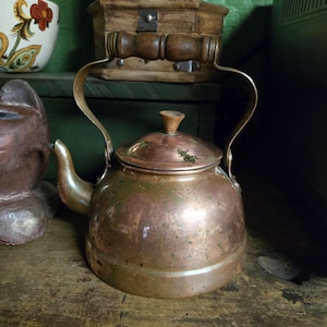 May include: A vintage copper kettle with a curved spout, a lid, and a wooden handle. The kettle has a tarnished, aged appearance, suggesting it is an antique. The handle is made of wood and is attached to the kettle with metal brackets.