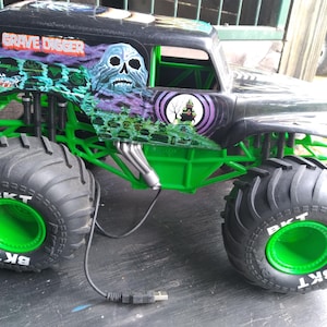 Pu&ograve; includere: Giocattolo di monster truck Grave Digger nero e verde con pneumatici grandi. Il camion presenta una grafica a teschio e le parole "Grave Digger". I pneumatici hanno le lettere "BKT" in bianco. Il camion ha un telaio e dettagli verdi.