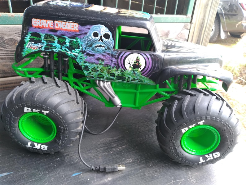 Pu&ograve; includere: Giocattolo di monster truck Grave Digger nero e verde con pneumatici grandi. Il camion presenta una grafica a teschio e le parole "Grave Digger". I pneumatici hanno le lettere "BKT" in bianco. Il camion ha un telaio e dettagli verdi.