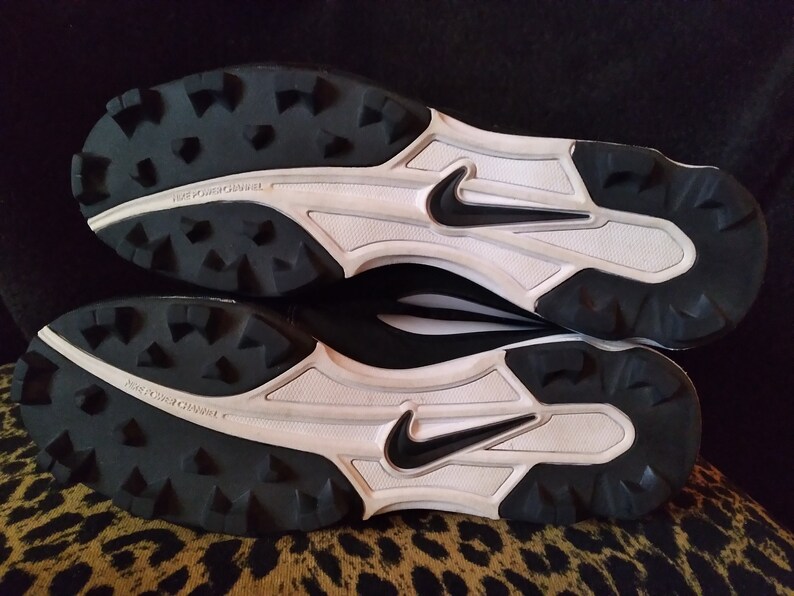 May include: Black and white baseball cleats with a Nike swoosh logo on the side. The cleats have a white midsole and a black outsole with a tread pattern.