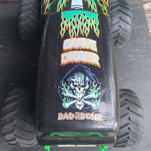 Pu&ograve; includere: Giocattolo di monster truck Grave Digger nero e verde con grandi pneumatici neri. Il camion presenta grafiche a fiamma e le frasi "Grave Digger" e "Bad to the Bone". Una maniglia verde &egrave; visibile sul davanti.