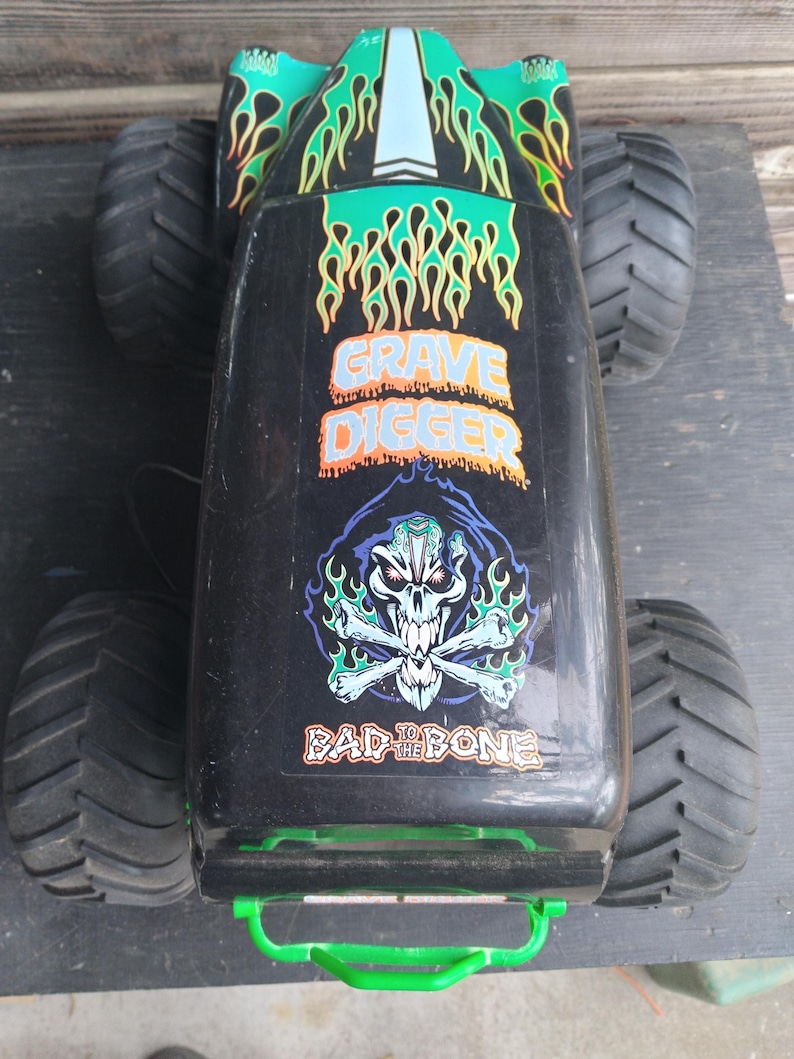 Pu&ograve; includere: Giocattolo di monster truck Grave Digger nero e verde con grandi pneumatici neri. Il camion presenta grafiche a fiamma e le frasi "Grave Digger" e "Bad to the Bone". Una maniglia verde &egrave; visibile sul davanti.