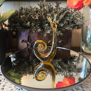 Könnte beinhalten: Eine dekorative bernsteinfarbene Glas-Schneckenfigur mit einem spiralförmigen Gehäuse und Fühlern. Die Schnecke steht auf einem Spiegel-Tablett, das ihre Form widerspiegelt. Der Hintergrund umfasst eine dekorative Pflanze und Blumen.