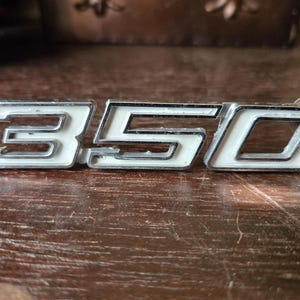 May include: A chrome and white "350" emblem, likely for a vehicle. The numbers are outlined in chrome with a white interior. The emblem is lying on a wooden surface.