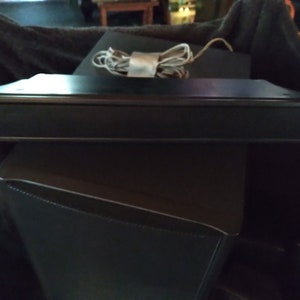 Puede incluir: Un dispositivo electr&oacute;nico rectangular negro con un borde plateado, posiblemente una barra de sonido o un altavoz. Un cable blanco y gris enrollado descansa encima. El dispositivo est&aacute; colocado sobre una superficie negra.