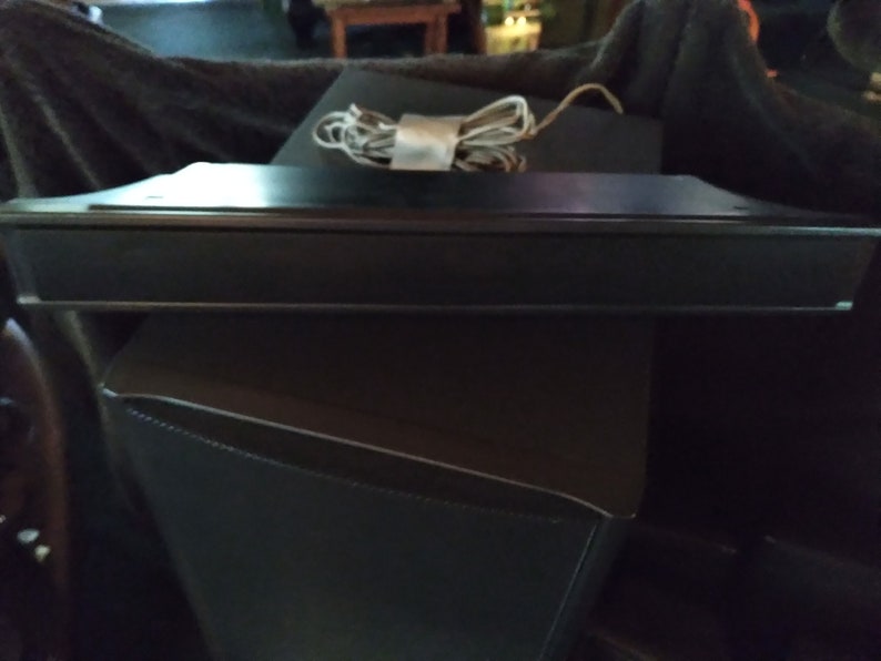 Puede incluir: Un dispositivo electr&oacute;nico rectangular negro con un borde plateado, posiblemente una barra de sonido o un altavoz. Un cable blanco y gris enrollado descansa encima. El dispositivo est&aacute; colocado sobre una superficie negra.