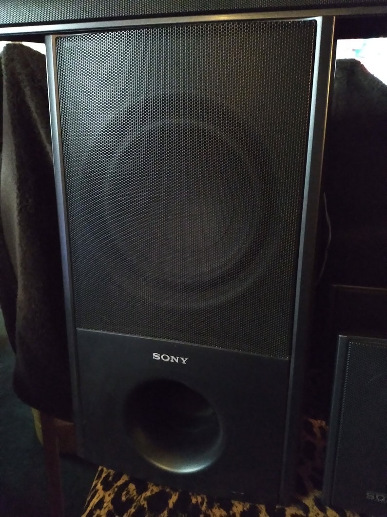 Puede incluir: Un altavoz Sony negro con un panel frontal de malla y un puerto circular en la parte inferior. El altavoz tiene forma rectangular y la marca "SONY" es visible en la parte delantera.