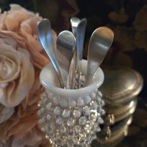 May include: A collection of small silver spoons in a white hobnail vase. The vase is decorated with raised, rounded glass beads. A stack of silver heart-shaped boxes is in the background, along with a bouquet of peach-colored flowers.