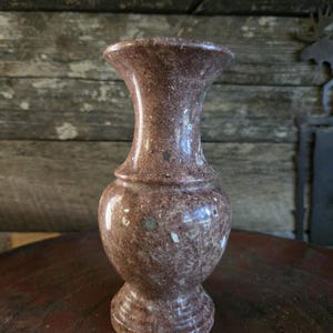 Puede incluir: Un jarrón decorativo de mármol marrón rojizo con una parte superior acampanada y un cuerpo redondeado. El jarrón tiene un patrón moteado y un acabado pulido, mostrando su textura de piedra natural. El jarrón mide unos 18 cm de alto.