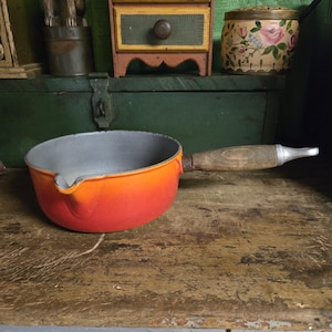 Könnte beinhalten: Ein orangefarbener und roter emaillierter Kochtopf mit Holzgriff. Der Innenraum ist grau. Die Pfanne hat einen Ausguss und steht auf einer Holzoberfläche. Der Griff ist mit einem silbernen Metallteil befestigt.