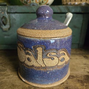 May include: A blue ceramic salsa jar with a lid and a small spoon. The word "Salsa" is carved into the side of the jar in a tan color. The jar has a speckled texture and a tan rim.
