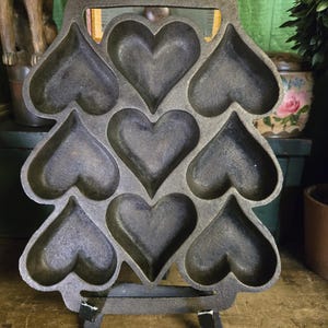 Pode incluir: Uma frigideira de ferro fundido cinza-escuro com sete cavidades em forma de coração dispostas num padrão vertical. O molde de metal pesado tem uma borda superior e inferior plana com uma superfície texturizada, concebido para cozinhar alimentos.