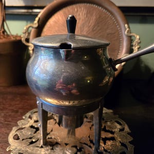 May include: A silver-toned metal pot with a rounded body, a lid featuring a black finial, and a side handle. The pot rests on a three-legged metal stand positioned atop a decorative, ornate silver-toned metal base with intricate floral cutouts.