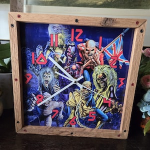 May include: Square wooden clock with a blue background featuring monster illustrations. The clock face has red numerals and white hands. The clock is framed in light brown wood with black and red accents.