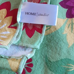 May include: A close-up of a floral patterned fabric in shades of green, red, yellow, and purple. The fabric has a tag that reads "HOME | studio". The floral design is a mix of large and small flowers.