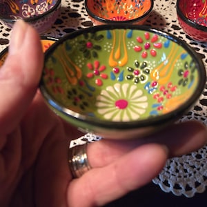 Könnte beinhalten: Kleine, handbemalte Keramikschale mit grünem Grund, verziert mit einem Blumenmuster in Weiß, Rosa, Gelb und Blau. Die Schale hat einen schwarzen Rand. Weitere Schalen mit ähnlichen Designs sind im Hintergrund zu sehen.