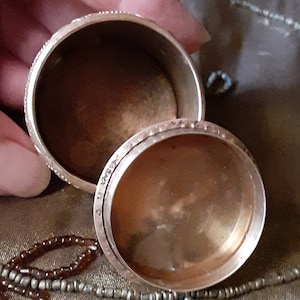 May include: A small, round, brass trinket box with a hinged lid. The box is open, revealing the inside, which is also made of brass. The box is sitting on a brown surface with beads.