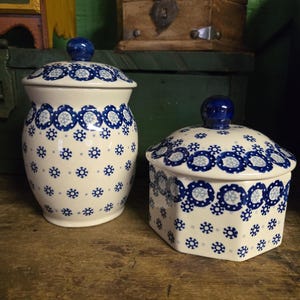 May include: Two ceramic jars with lids. The jars are white with a blue floral pattern and blue knob handles. One jar is round, and the other is hexagonal. The jars are on a wooden surface.