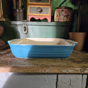 Puede incluir: Una fuente rectangular de cerámica azul claro con interior crema. La fuente tiene una forma ligeramente curvada y pequeñas asas. Se encuentra sobre una superficie de madera rústica y desgastada.