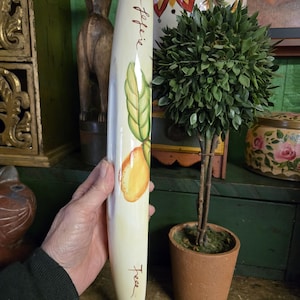 May include: A cream-coloured rolling pin with a painted design of a lemon and green leaves. The words "Plum feferie" and "Tees" are written on the pin in brown script. The rolling pin is held in a person's hand.