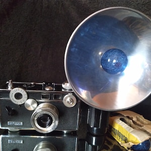 May include: A vintage black and silver rangefinder camera with a large, circular metallic flash attachment featuring a blue bulb. The camera body has silver dials and a lens with visible text. A small yellow box labeled flashbulbs sits nearby.