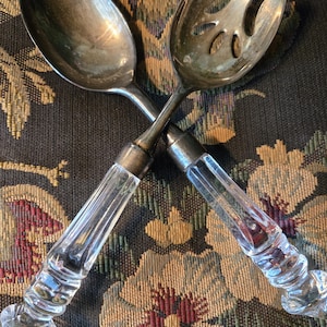 May include: A pair of silver-toned salad serving utensils consisting of a solid spoon and a slotted fork. Both pieces feature clear, faceted glass handles with a decorative, multi-tiered design and metallic ferrules connecting the metal heads.