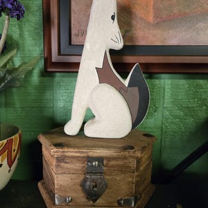May include: A white wooden fox figurine with brown and black details sits on a small, hexagonal wooden box with metal fittings. The fox is in a seated pose and has a stylized design. The box has a metal clasp and hinges.