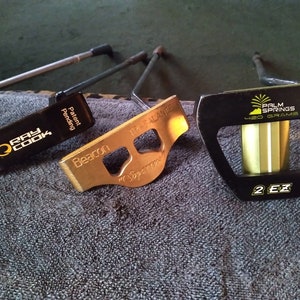 Könnte beinhalten: Drei verschiedene Golf-Putter. Der erste ist schwarz mit den Worten "Ray Cook" und "Patent Pending". Der zweite ist golden mit den Worten "Beacon" und "TR-SALANTIE". Der dritte ist schwarz mit den Worten "Palm Springs" und "420 Grams" und "2 EZ".