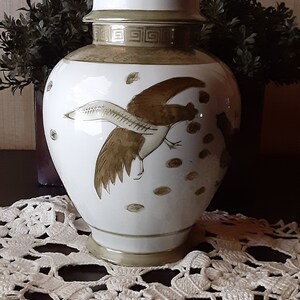 May include: A white porcelain ginger jar with a gold lid and a brown bird design. The jar is sitting on a white crocheted doily.