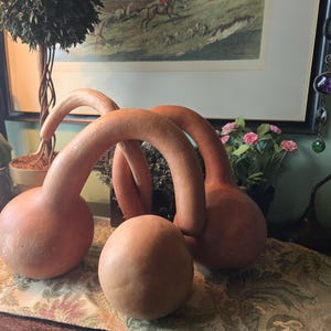 May include: Two long-necked, tan-coloured gourds with rounded bases are arched and intertwined to form a bridge-like structure. Positioned in front of the arched gourds is a single, smaller, smooth, tan-coloured spherical gourd.