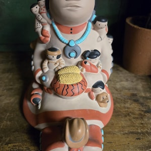 May include: A ceramic figurine of a woman with multiple small figures attached. The figurine is painted in cream, red, and turquoise. The woman wears a turquoise necklace and holds a basket. A cowboy hat is at the bottom.