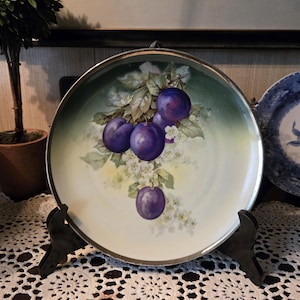 May include: A decorative plate with a silver rim, featuring a hand-painted design of purple plums, white blossoms, and green leaves. The plate is displayed on a black stand, showcasing its vintage charm and artistic detail.