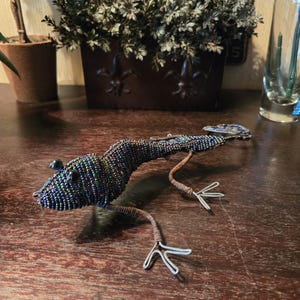 以下が含まれることがあります： 青、緑、紫の色合いの虹色のビーズで作られたビーズのトカゲの彫刻。 トカゲはワイヤー製の足と尾を持ち、木製の表面に展示されています。 この彫刻は装飾品です。