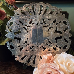 Könnte beinhalten: Ein silberfarbener Metalluntersetzer mit einem komplexen durchbrochenen Design und geprägten Blumen- und Blattmotiven. Das kreisförmige Stück besteht aus zwei symmetrischen Hälften, die in der Mitte verbunden sind und feine Schnörkel sowie gewellte Ränder aufweisen.
