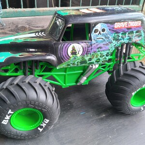 Pu&ograve; includere: Un giocattolo di monster truck nero e verde con le parole "Grave Digger" e una grafica a teschio sul lato. Il camion ha grandi pneumatici neri con cerchi verdi e le lettere "BKT" sugli pneumatici.
