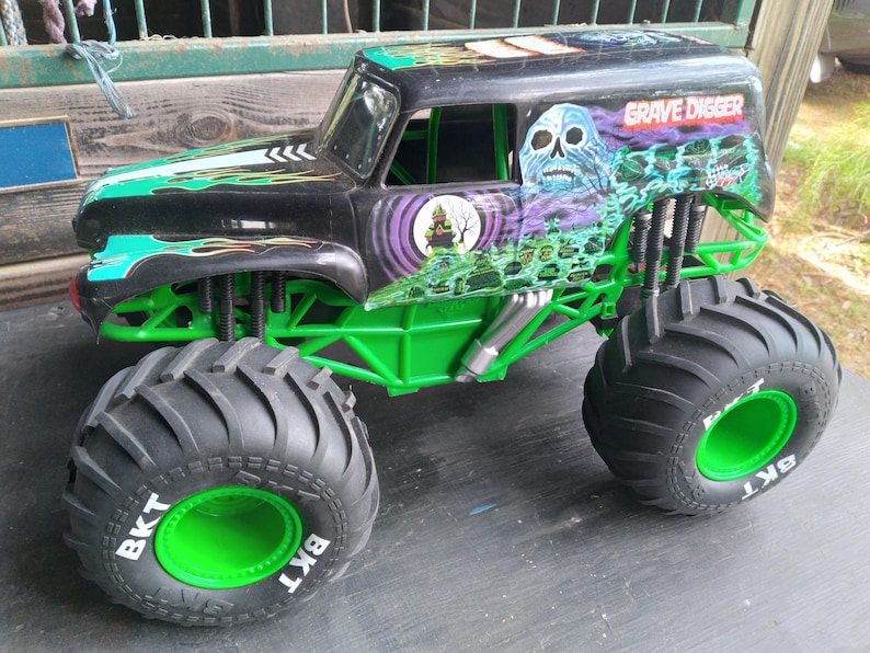 Pu&ograve; includere: Un giocattolo di monster truck nero e verde con le parole "Grave Digger" e una grafica a teschio sul lato. Il camion ha grandi pneumatici neri con cerchi verdi e le lettere "BKT" sugli pneumatici.