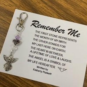 May include: A white card with the text "Remember Me" and a poem about the meaning of each charm on a keychain. The keychain has a silver angel charm, a purple crystal charm, a silver heart charm, and a silver bead charm.