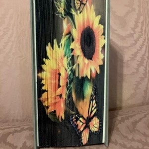 May include: A book art sculpture with a black background and a design of sunflowers and butterflies. The sunflowers are yellow and orange with brown centers. The butterflies are orange and black.