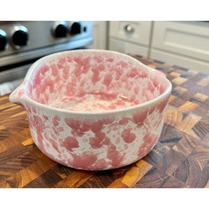 May include: A small, pink and white speckled ceramic bowl with a rounded shape and a pouring spout. The bowl is sitting on a wooden cutting board.